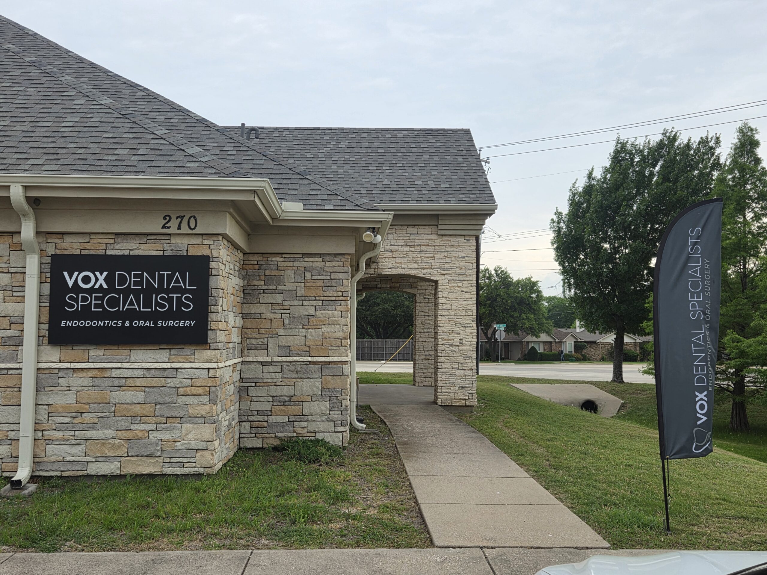 Picture of the outside of the office. Sidewalk on the right, black sign on the left with our name. Door at the end of the sidewalk on the left
